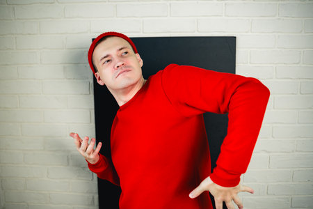 A man in a red hoodie and hat dances against a white brick wall.