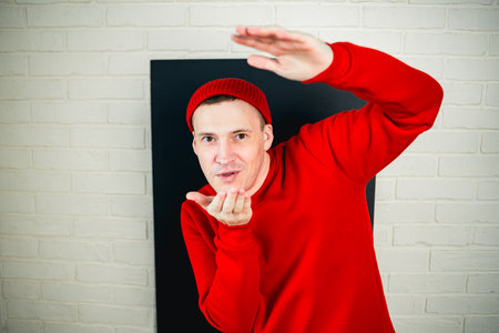 A man in a red hoodie and hat dances against a white brick wall.