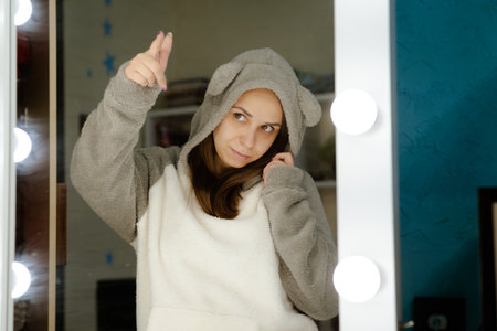 A young woman dances energetically in front of a mirror in her bedroom. Shes wearing a gray hoodie and smiling as she moves.の写真素材