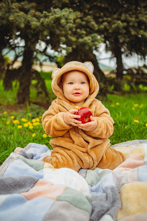 Cute baby girl in plush suit with red apple in hands sitting on blanket in green grass