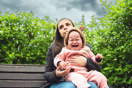 A tired mother sits on a park bench with her baby who is crying hysterically. She looks exhausted and overwhelmed.の写真素材