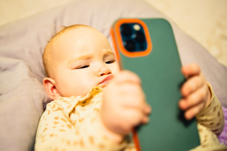 Baby is holding a cell phone in its hand, looking at the screen with curiosity, showing the contrast...