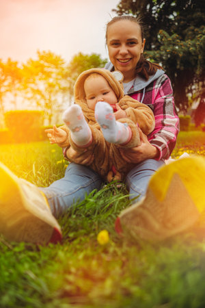 A mother and her baby enjoy a beautiful spring day in the park. They are sitting on a blanket,...