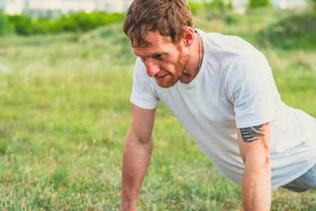 Male Athlete Performing Push-Ups on Grass in Sunny Outdoor Park. Man athlete is engaged in a robust...