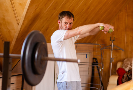 A man is lifting green dumbbells in his home. He is wearing a white t-shirt and has a serious expression on his face.の写真素材