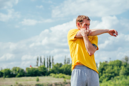 A man in a yellow t-shirt stretches his arm while standing outside on a sunny day. He is stretching his arm and shoulder muscles.の写真素材
