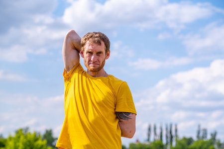 A man in a yellow t-shirt stretches his arm while standing outside on a sunny day. He is stretching...