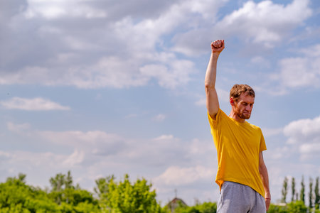 A man in a yellow t-shirt warms up his arm by stretching it above his head. He is outdoors with...