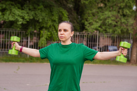 A woman in a green t-shirt performs a dumbbell shoulder press exercise during an outdoor workout...