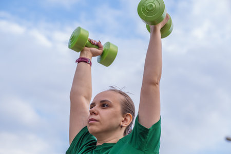 A woman in a green t-shirt performs a dumbbell shoulder press exercise during an outdoor workout...