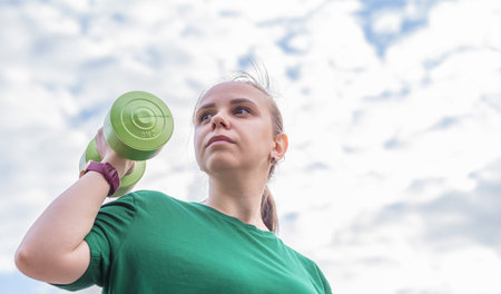 A woman in a green t-shirt performs a dumbbell shoulder press exercise during an outdoor workout...