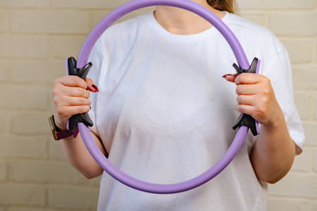 A woman in a white shirt is using a pilates ring for an exercise, standing in front of a white brick...