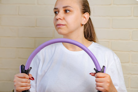 A woman in a white shirt is using a pilates ring for an exercise, standing in front of a white brick wallの写真素材