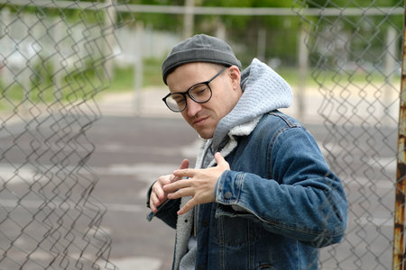 A Man with glasses in a Denim Jacket Dances Near a Chain Link Fence.