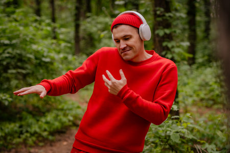 A man in a red sweater and a red beanie is dancing in a forest. He is wearing headphones and seems to be enjoying the music. The trees and greenery behind him create a beautiful and peaceful backdropの写真素材