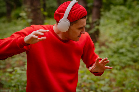 A man in a red sweater and a red beanie is dancing in a forest. He is wearing headphones and seems to be enjoying the music. The trees and greenery behind him create a beautiful and peaceful backdropの写真素材