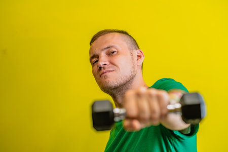 A man in a green t-shirt stands against a bright yellow background, his arm raised as he lifts a dumbbellの写真素材