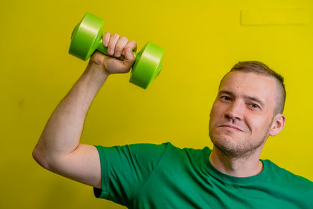 A man in a green t-shirt stands against a bright yellow background, his arm raised as he lifts a dumbbellの写真素材