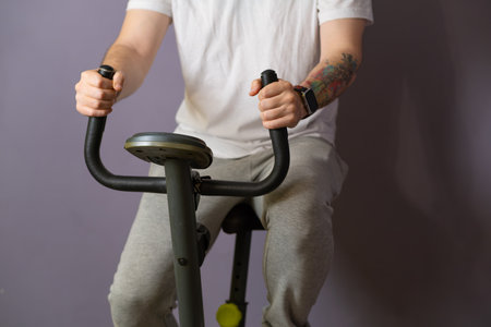 A close-up view of a mans hands gripping the handlebars of a stationary exercise bike. He is wearing a white t-shirt and his focus is intense as he pedals for a good workoutの写真素材