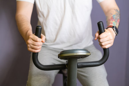 A close-up view of a mans hands gripping the handlebars of a stationary exercise bike. He is wearing...