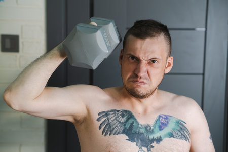 A shirtless man with a tattoo of a bird on his chest lifts a gray dumbbell with a determined expression on his faceの写真素材
