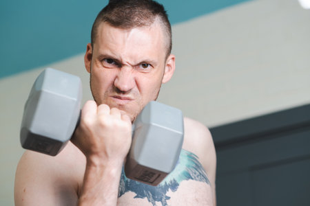 A shirtless man with a tattoo of a bird on his chest lifts a gray dumbbell with a determined expression on his faceの写真素材