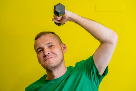 A man in a green t-shirt stands against a bright yellow background, his arm raised as he lifts a dumbbellの写真素材