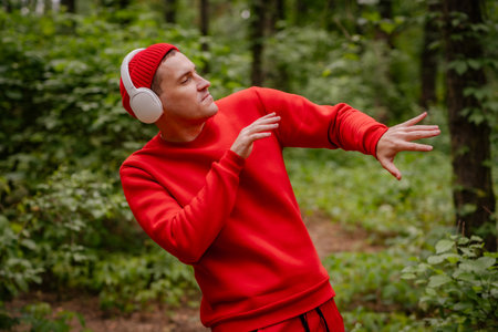 A man in a red sweater and a red beanie is dancing in a forest. He is wearing headphones and seems to be enjoying the music. The trees and greenery behind him create a beautiful and peaceful backdropの写真素材