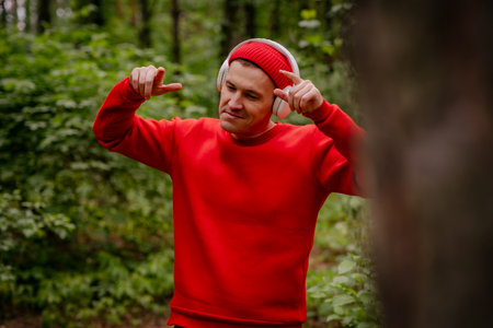 A man in a red sweater and a red beanie is dancing in a forest. He is wearing headphones and seems to be enjoying the music. The trees and greenery behind him create a beautiful and peaceful backdropの写真素材