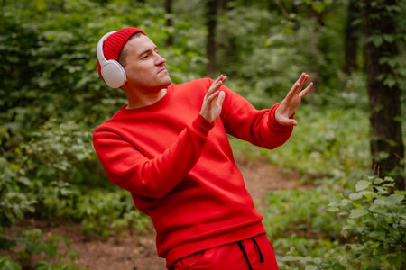 A man in a red sweater and a red beanie is dancing in a forest. He is wearing headphones and seems to be enjoying the music. The trees and greenery behind him create a beautiful and peaceful backdropの写真素材