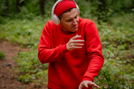 A man in a red sweater and a red beanie is dancing in a forest. He is wearing headphones and seems to be enjoying the music. The trees and greenery behind him create a beautiful and peaceful backdropの写真素材