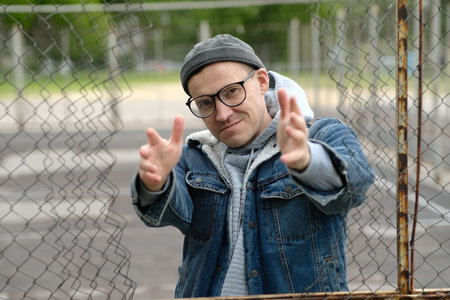 A Man with glasses in a Denim Jacket Dances Near a Chain Link Fence.