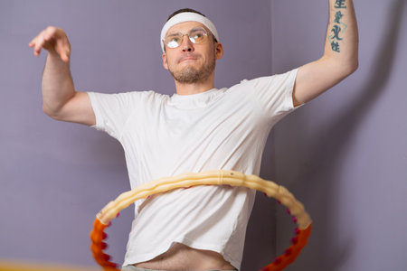 Man In White T-Shirt And Headband Using A Weighted Hula Hoop For Exercise In Front Of Purple Wall.