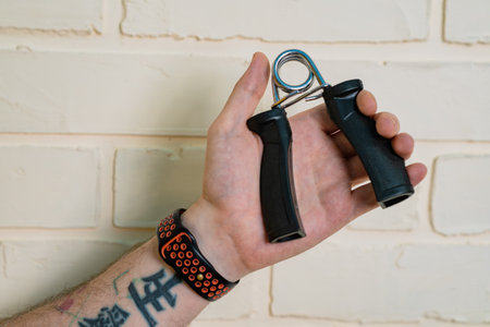 A Mans Arm With A Tattoo Flexes His Hand With A Gripping Device In Front Of A White Brick Wallの写真素材