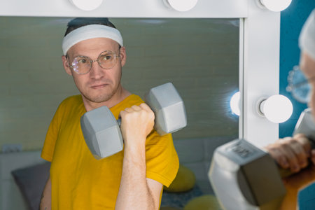 A man wearing a headband and glasses lifts a dumbbell in front of a vanity mirror.の写真素材