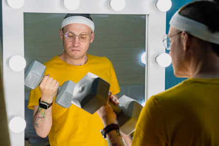 A man wearing a headband and glasses lifts a dumbbell in front of a vanity mirror.の写真素材
