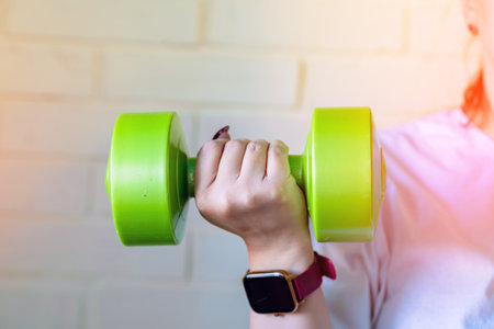 A woman lifts a green dumbbell with her right arm, while wearing a smartwatch and a white shirt.の写真素材