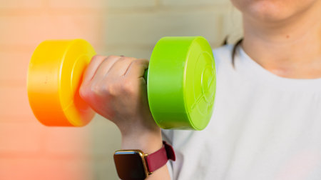 A woman lifts a green dumbbell with her right arm, while wearing a smartwatch and a white shirt.の写真素材