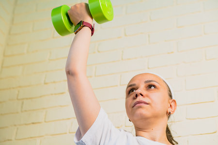 A woman lifts a green dumbbell with her right arm, while wearing a smartwatch and a white shirt.
