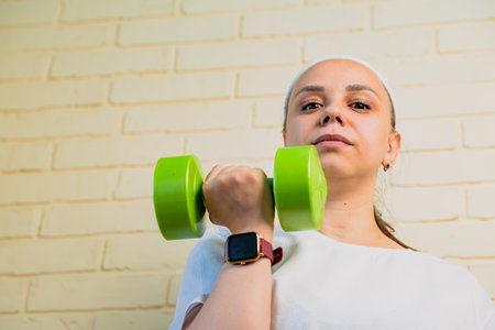 A woman lifts a green dumbbell with her right arm, while wearing a smartwatch and a white shirt.