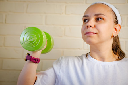 A woman lifts a green dumbbell with her right arm, while wearing a smartwatch and a white shirt.