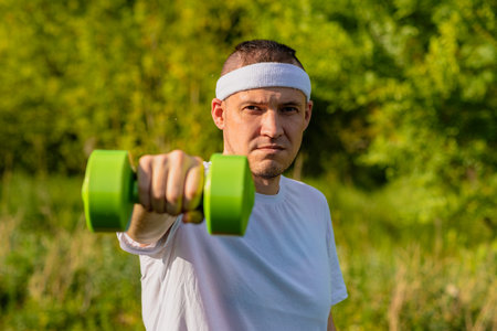 A man in a white t-shirt and headband lifts a green dumbbell outside on a sunny day.の写真素材