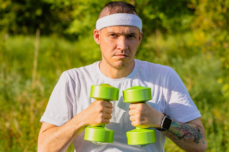 A man in a white t-shirt and headband lifts a green dumbbell outside on a sunny day.