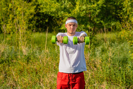 A man in a white t-shirt and headband lifts a green dumbbell outside on a sunny day.の写真素材