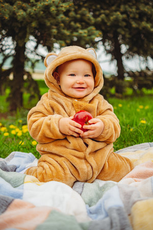 Cute baby girl in plush suit with red apple in hands sitting on blanket in green grassの写真素材
