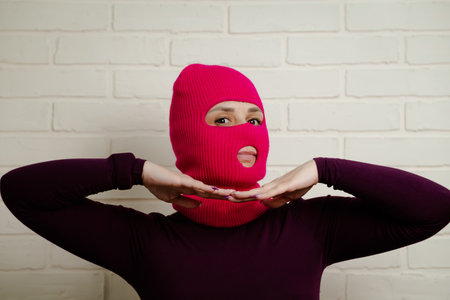 A woman wearing a bright pink balaclava dances energetically, striking unique poses with her hands against a clean, white brick wall background.の写真素材