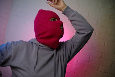 Individual in a pink balaclava poses against a colorful backdrop, making a gesture.