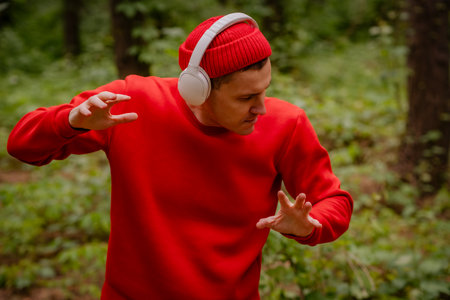 A man in a red sweater and a red beanie is dancing in a forest. He is wearing headphones and seems to be enjoying the music. The trees and greenery behind him create a beautiful and peaceful backdropの写真素材