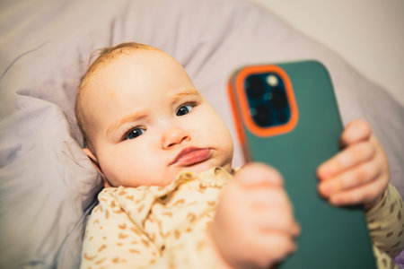 Baby is holding a cell phone in its hand, looking at the screen with curiosity, showing the contrast...