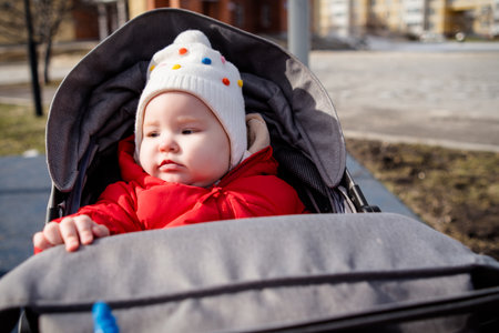 Baby in a Red Jacket and White Hat Seated in a Stroller on a Sunny Day. A baby dressed warmly looks...